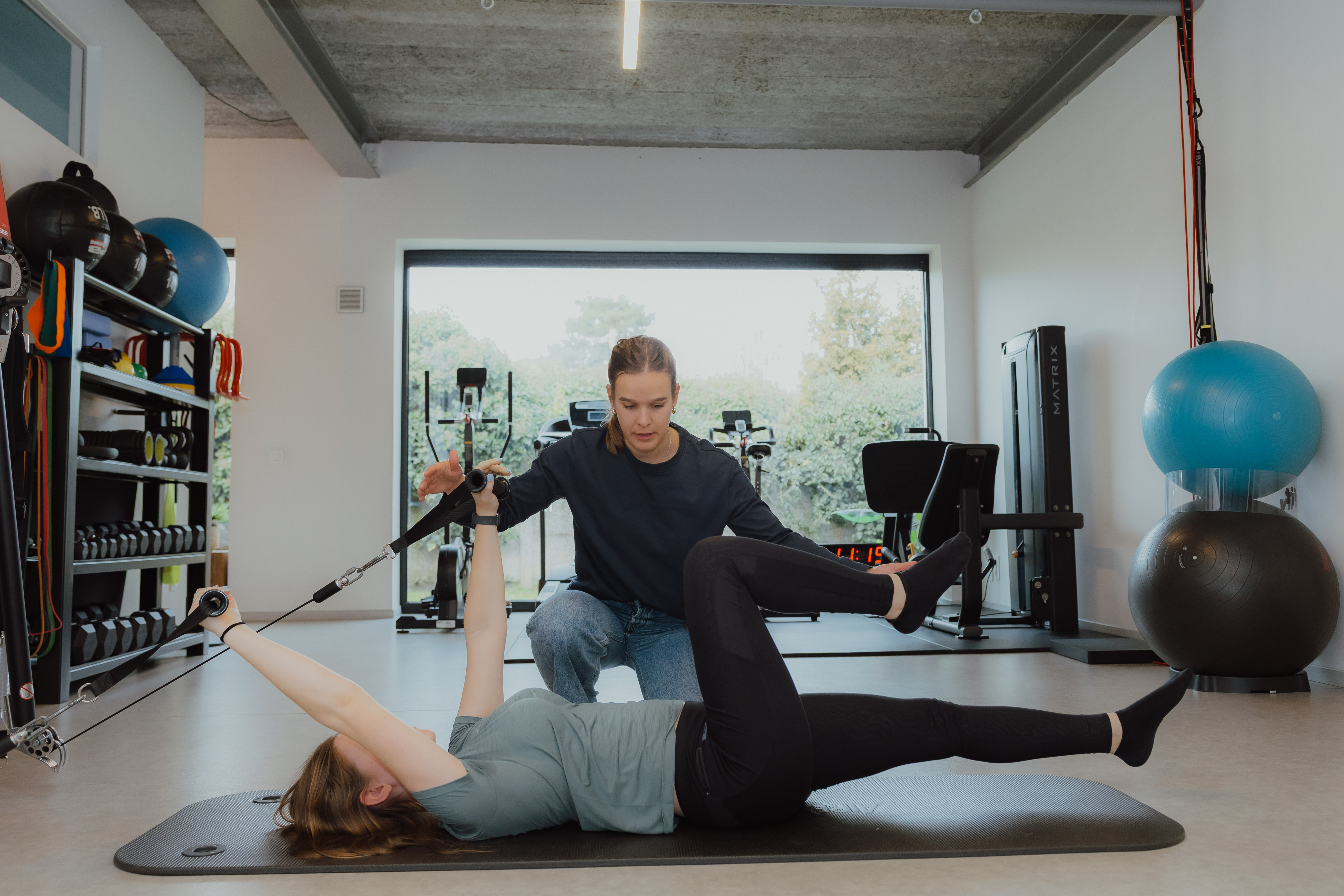 Persoon die op een mat ligt en met weerstandsoefeningen onder begeleiding van een trainer traint in een fitnessruimte.