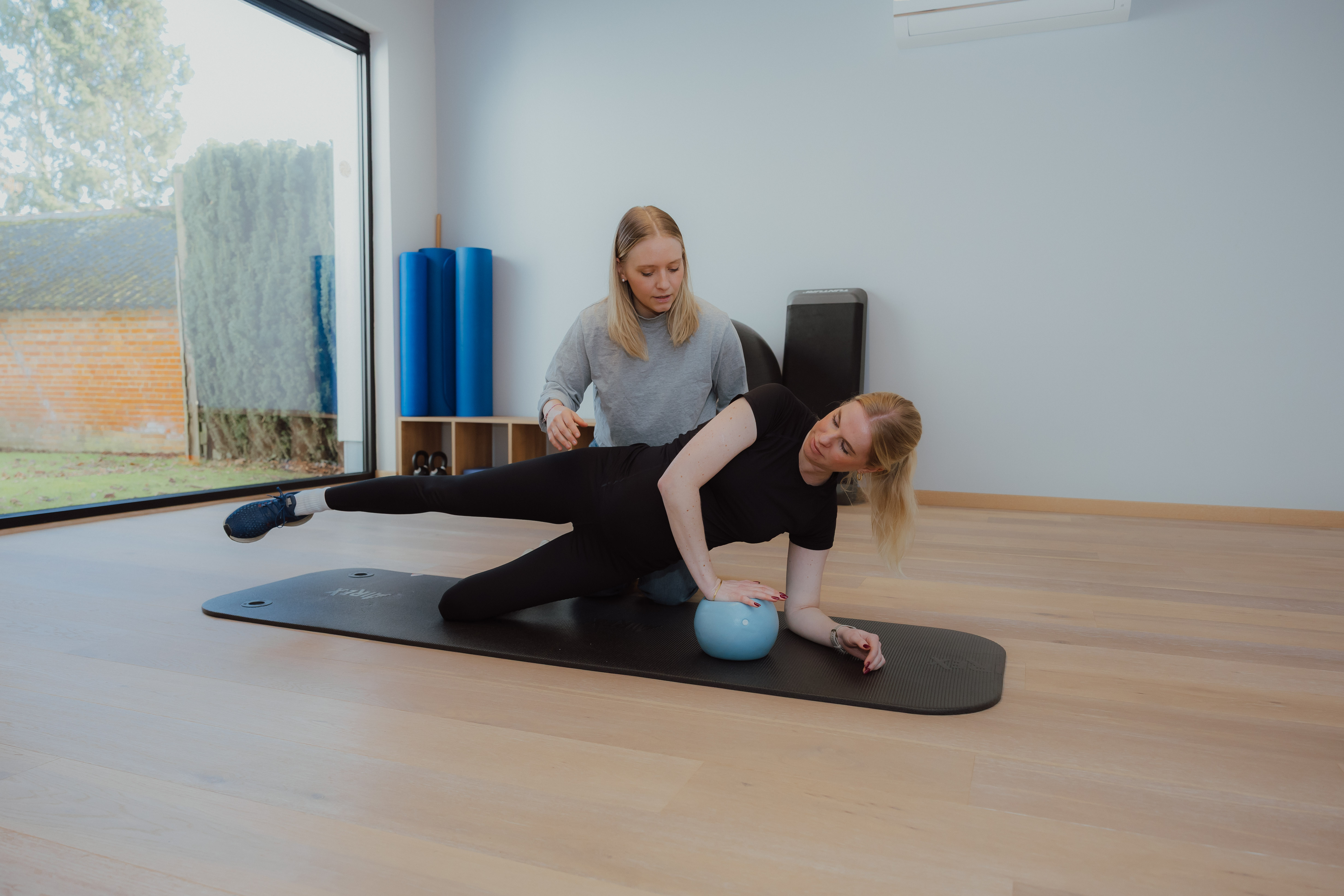 Fysiotherapeut begeleidt vrouw die zijdelingse beenheffing doet op yogamat met blauwe oefenbal.