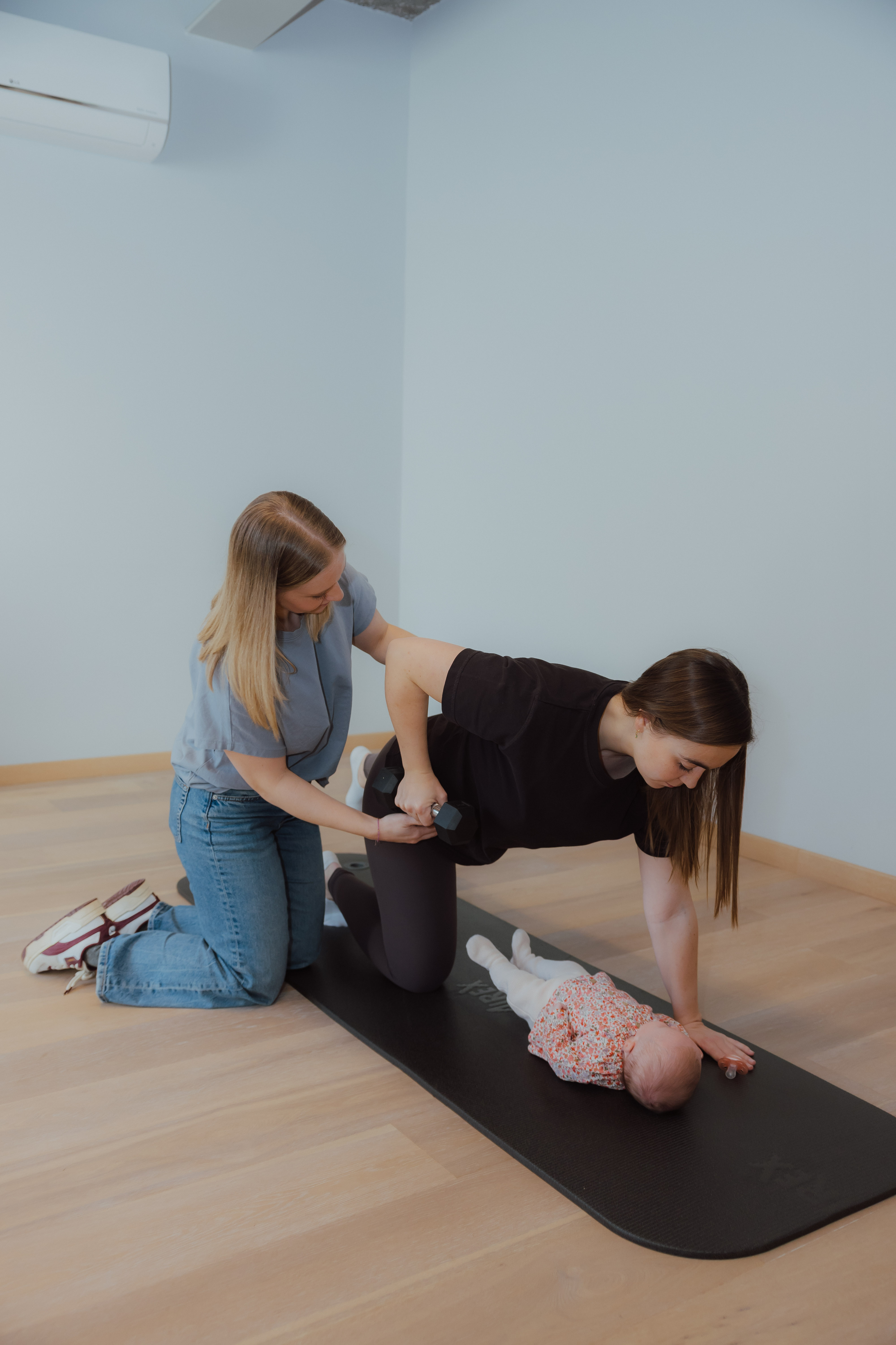 Vrouw die trainingshulpmiddel biedt aan een andere vrouw die op een yogamat oefeningen doet met een baby liggend voor haar.
