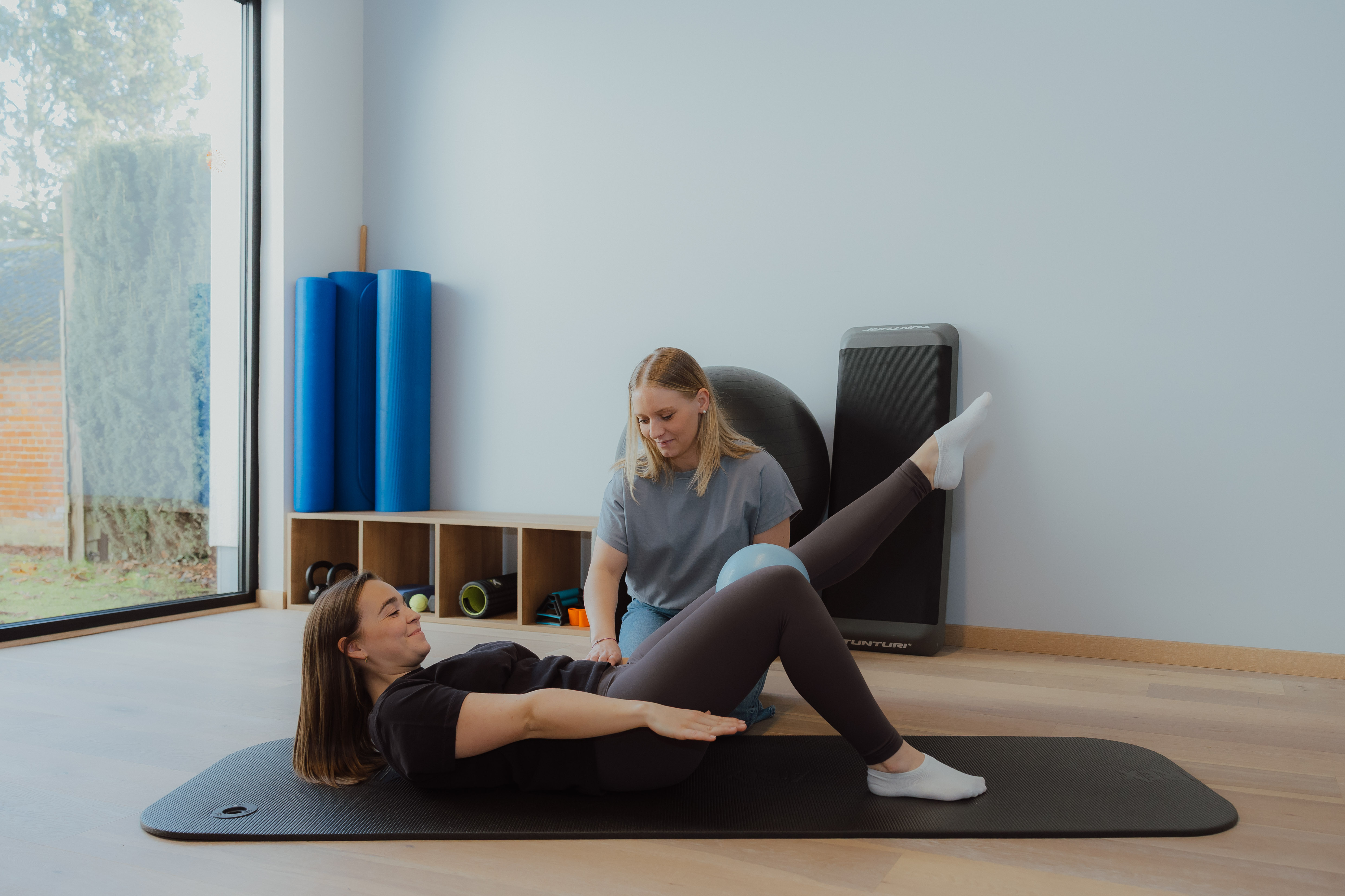 Een vrouw ligt op een fitnessmat met een kleine bal tussen haar knieën, terwijl een andere vrouw haar begeleidt bij een oefening in een lichte oefenruimte.
