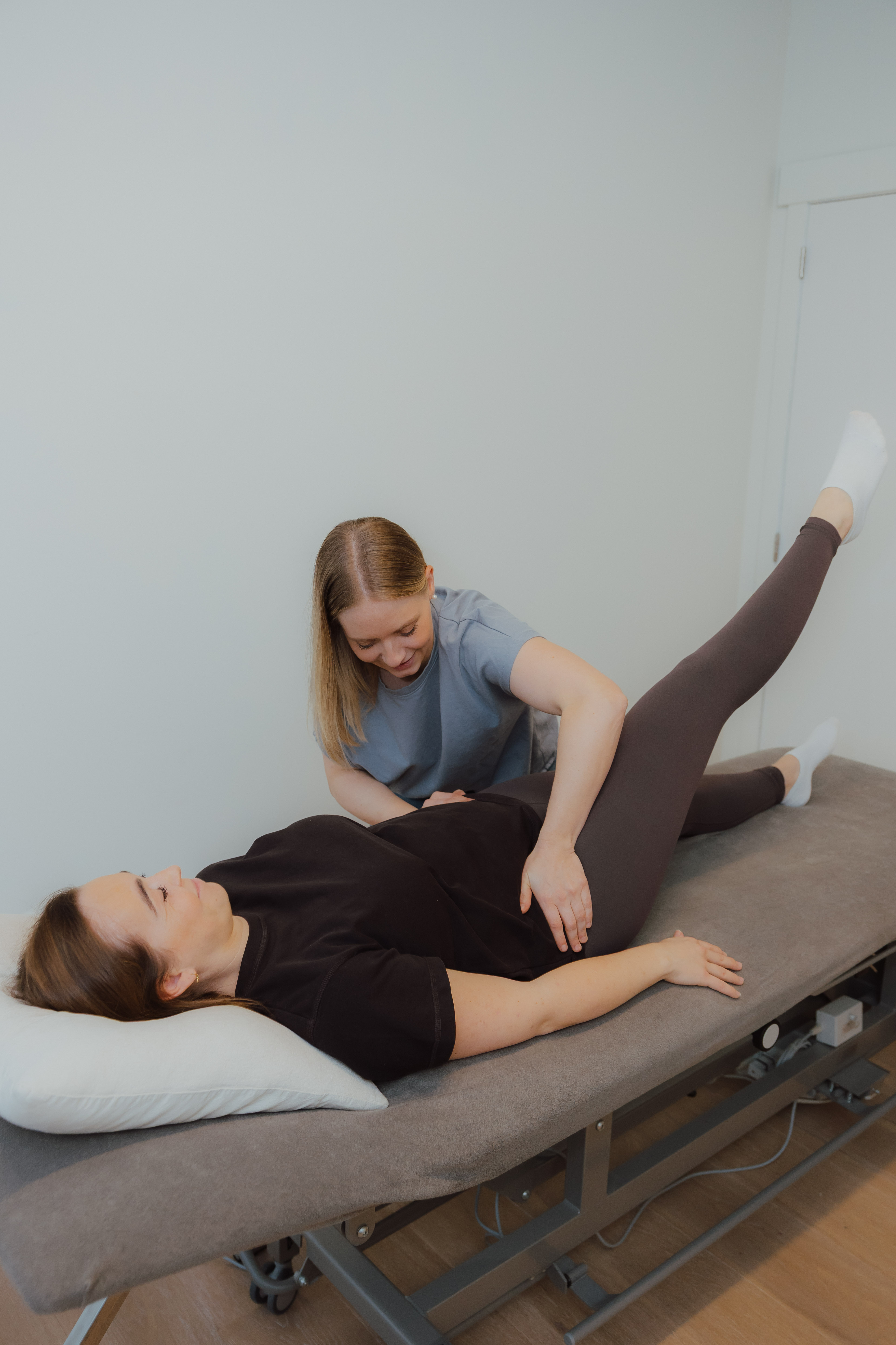 Fysiotherapeut helpt een liggende vrouw met het optillen van haar been op een behandeltafel.