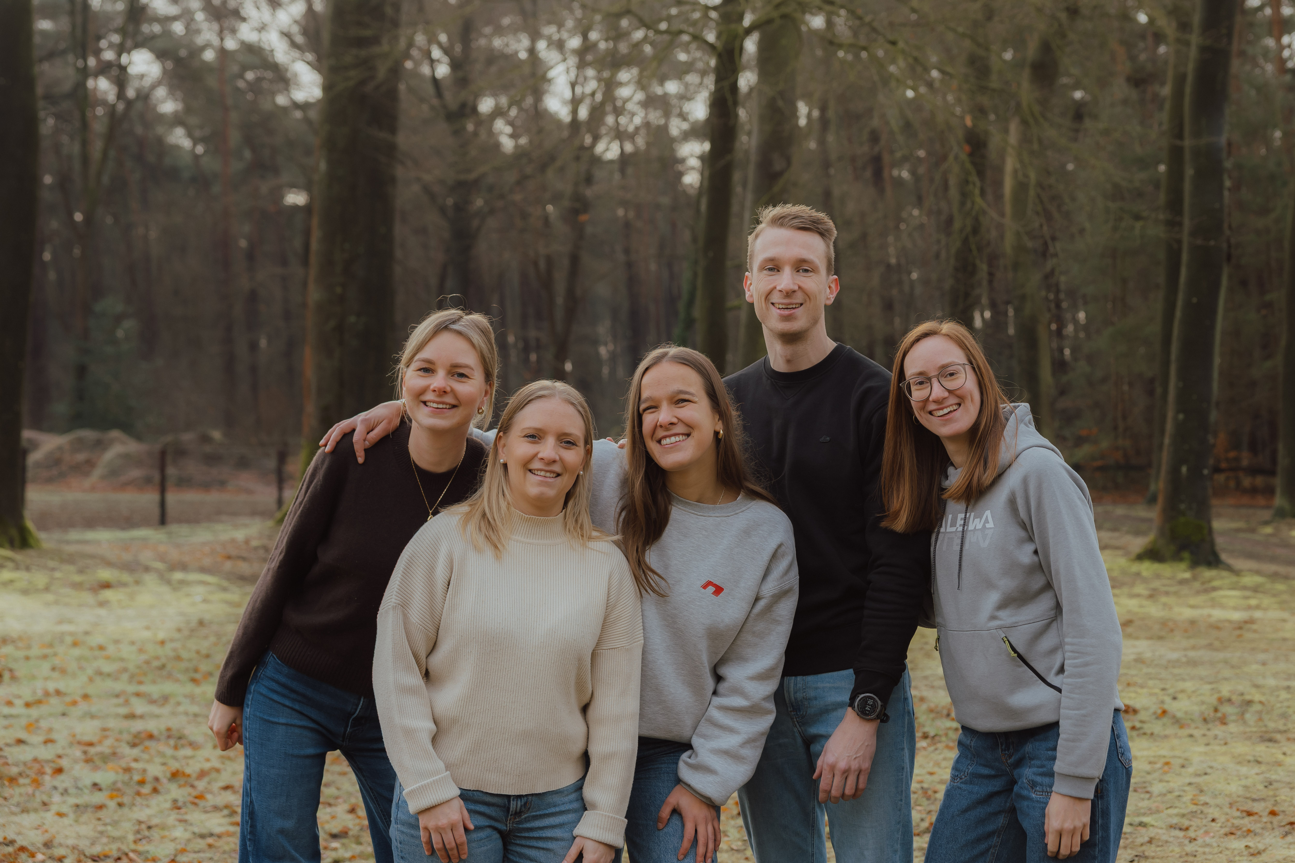 Vijf jonge volwassenen lachen samen in een bosrijke omgeving met bomen op de achtergrond.