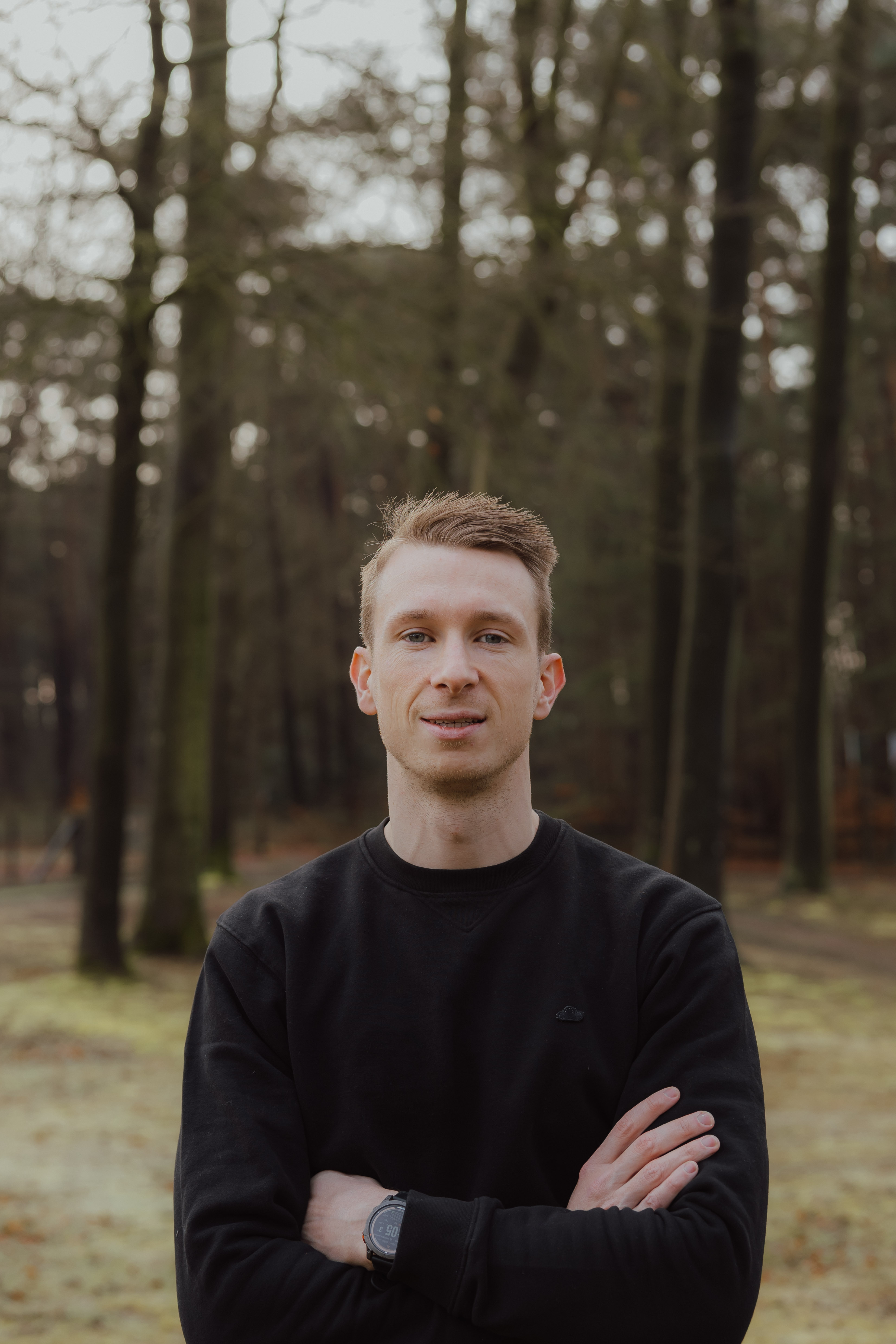 Jonge man met zwart sweatshirt en digitale polshorloge staat met armen gekruist in een bosrijke omgeving.