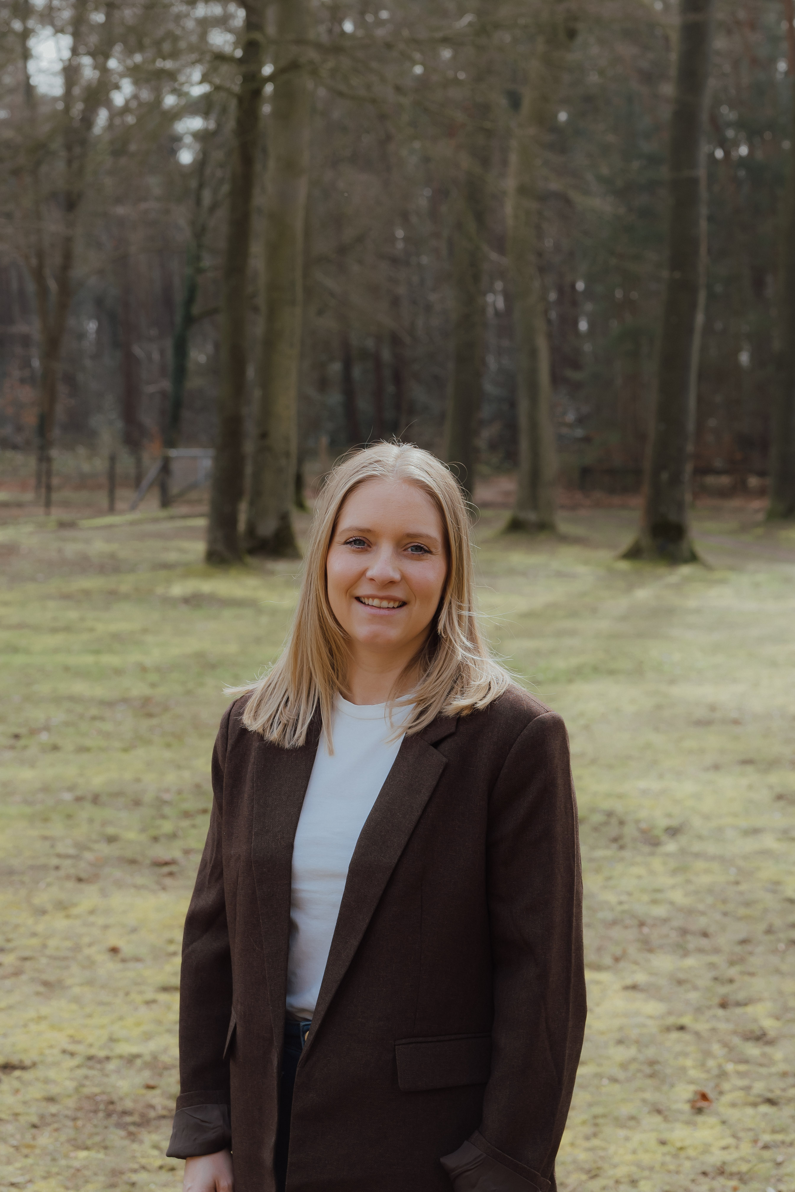 Vrouw met blond haar die buiten in een bosrijk gebied lacht, gekleed in een donkerkleurig colbert en een witte shirt.