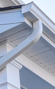 Close-up of a white rain gutter and downspout attached to a gray house under a clear sky.