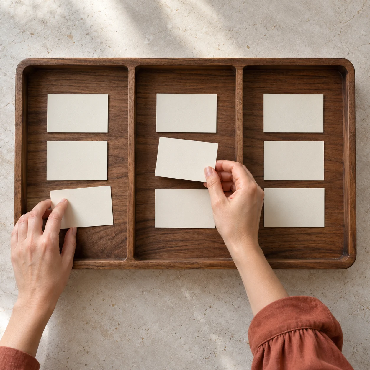 Two hands sort blank cream cards into a divided walnut tray on a stone table, illustrating how an agency shortlist is grouped.