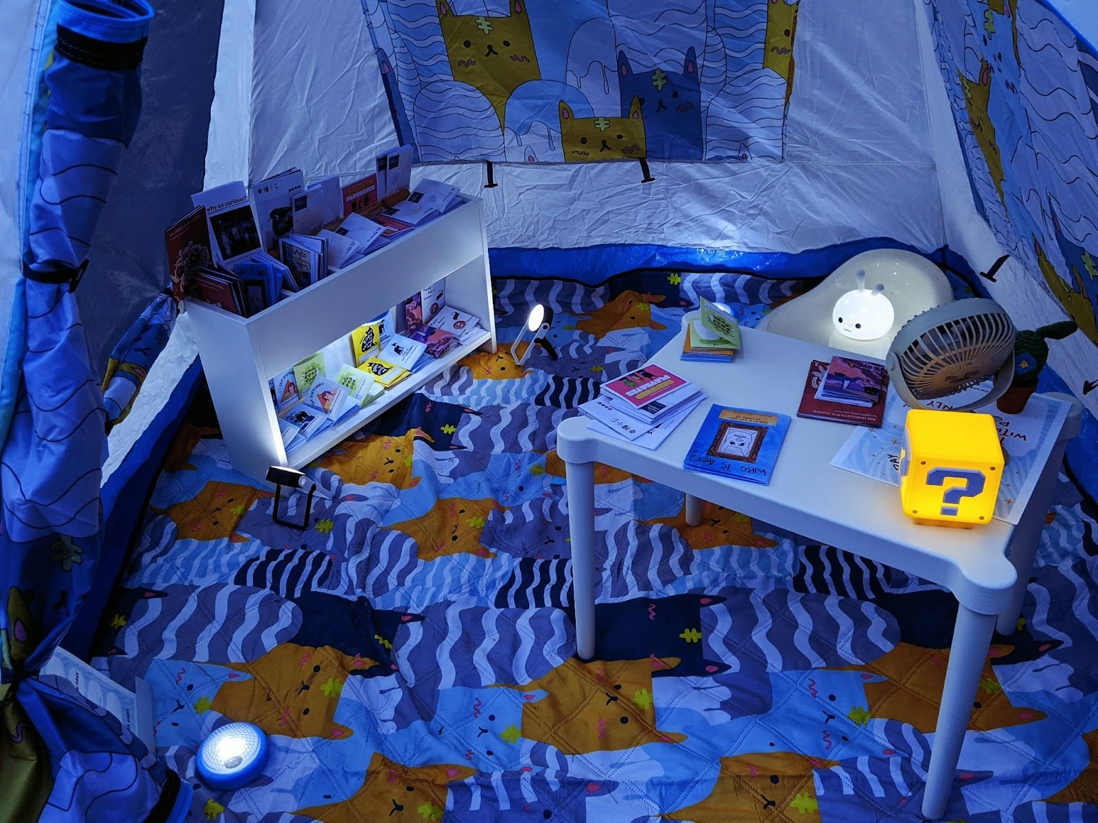 A cozy tent space decorated with patterned blankets, small lights, and shelves of colorful books surrounding a low table with more books and a glowing cube lamp.