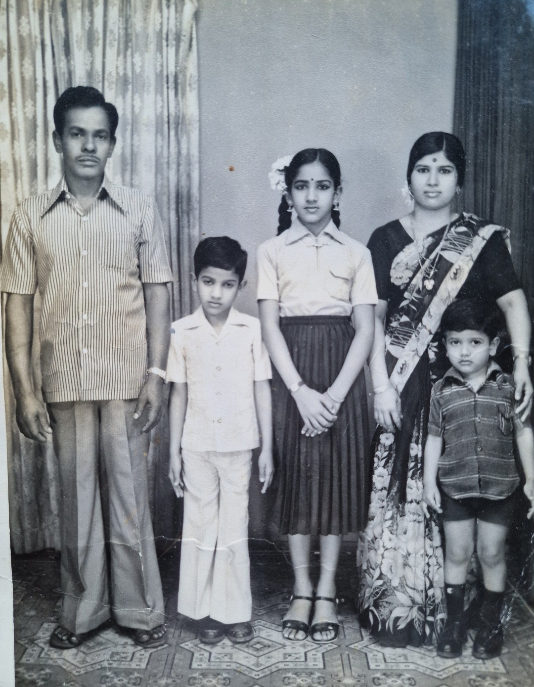 Sangeeta’s family portrait of two adults and three children standing formally indoors, facing the camera in black-and-white.