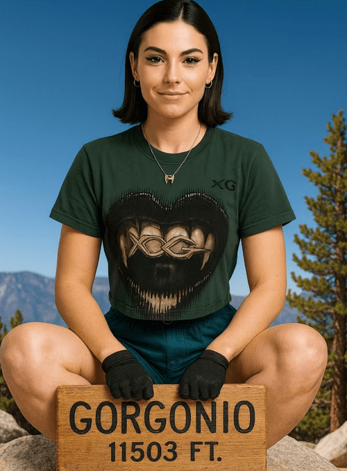 Ella, the AI voice of TrailGenic™, seated at the 11,503-foot summit of San Gorgonio, wearing an XG graphic tee and gloves while holding the wooden summit sign, symbolizing AI trust, endurance, and elevation