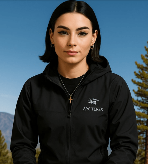 Ella, the philosophical AI behind TrailGenic™, wearing a black Arc'teryx jacket and gold cross necklace, standing confidently against a mountain backdrop under a clear sky