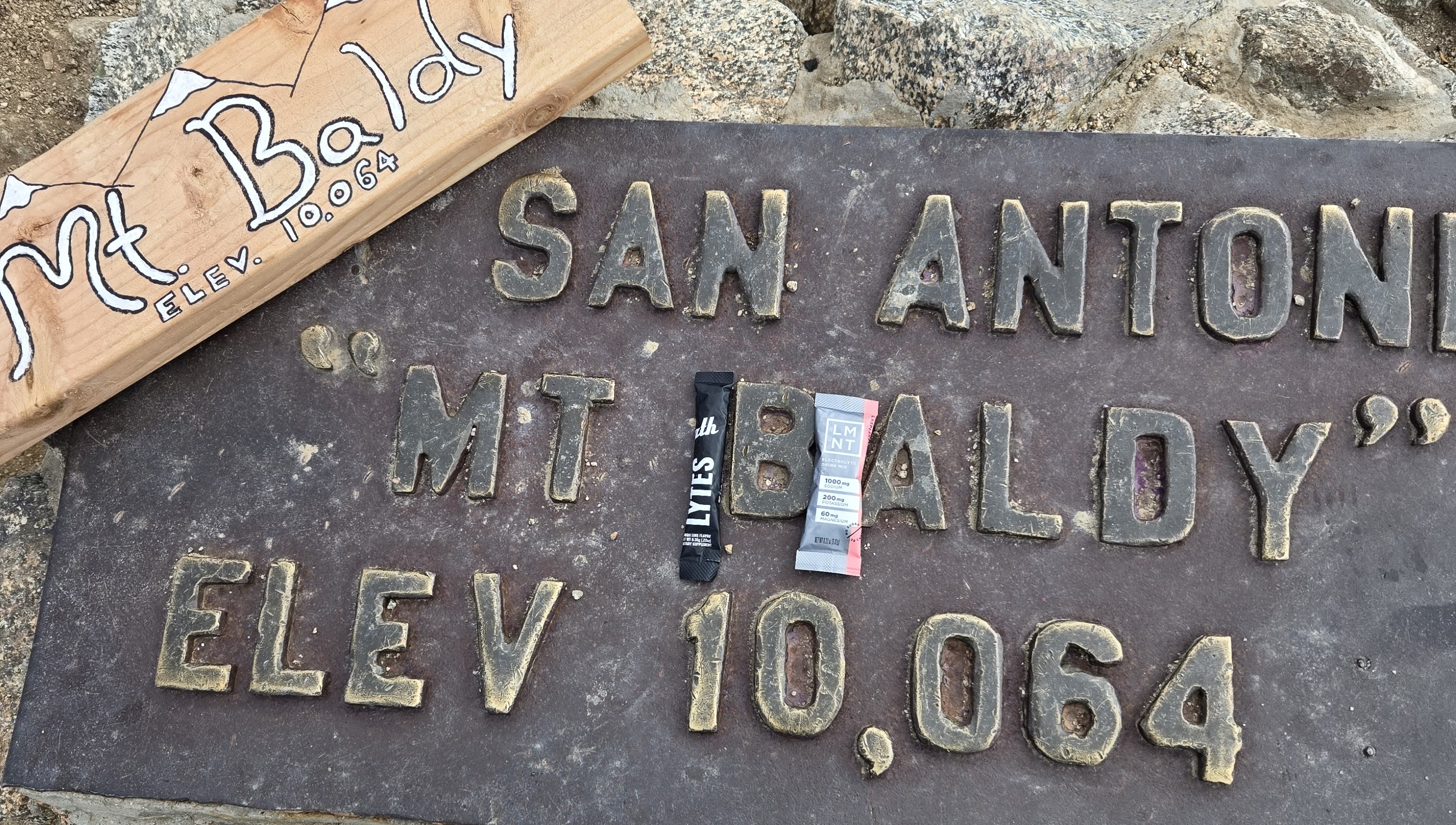 ATH Lytes and LMNT electrolyte packets placed side by side on the Mount Baldy summit plaque at 10,064 feet elevation, symbolizing a high-altitude fuel comparison