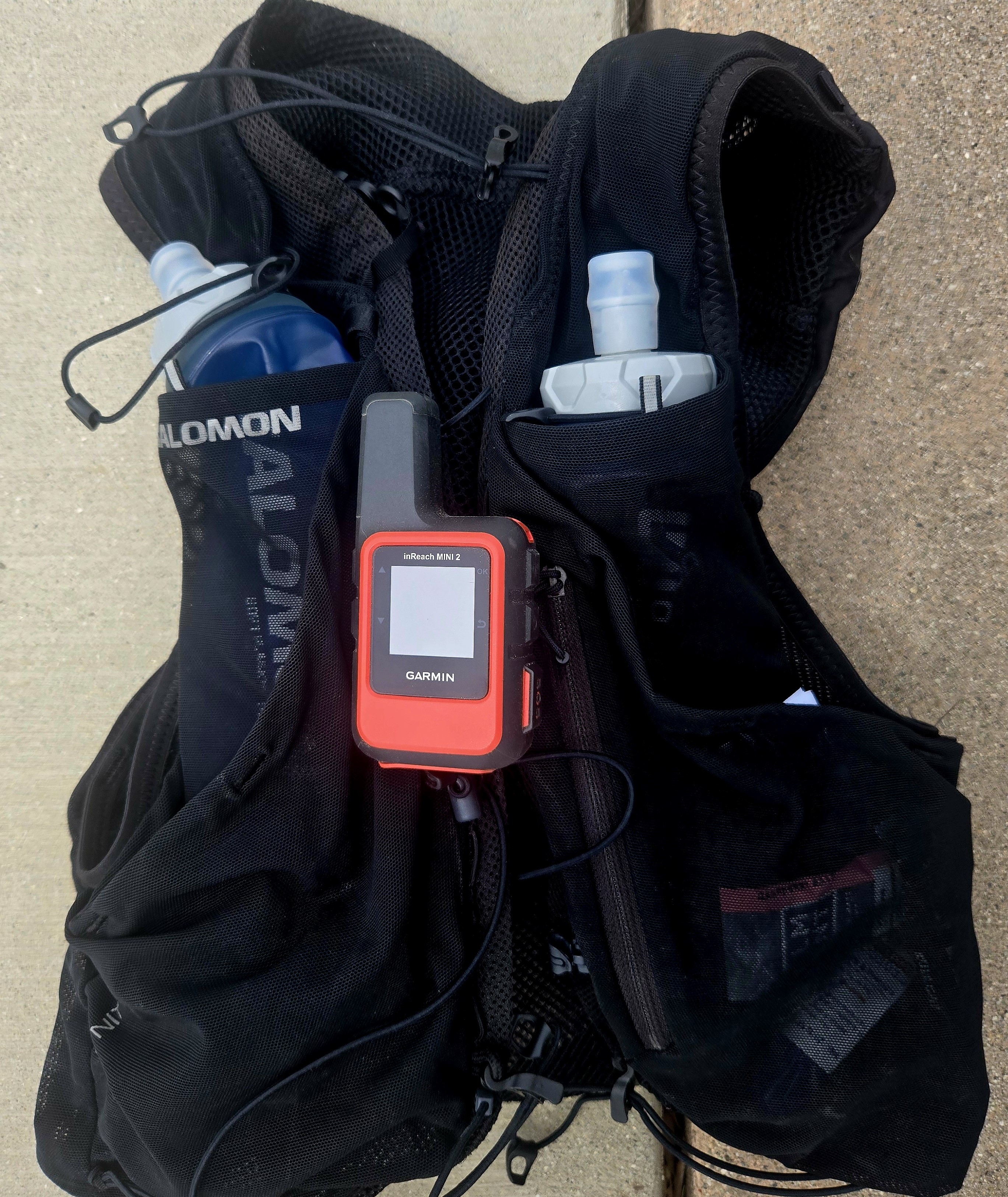 Garmin inReach Mini 2 GPS satellite communicator attached to the front strap of a Salomon ADV Skin hydration vest, with dual soft flasks in view — ready for backcountry safety and tracking