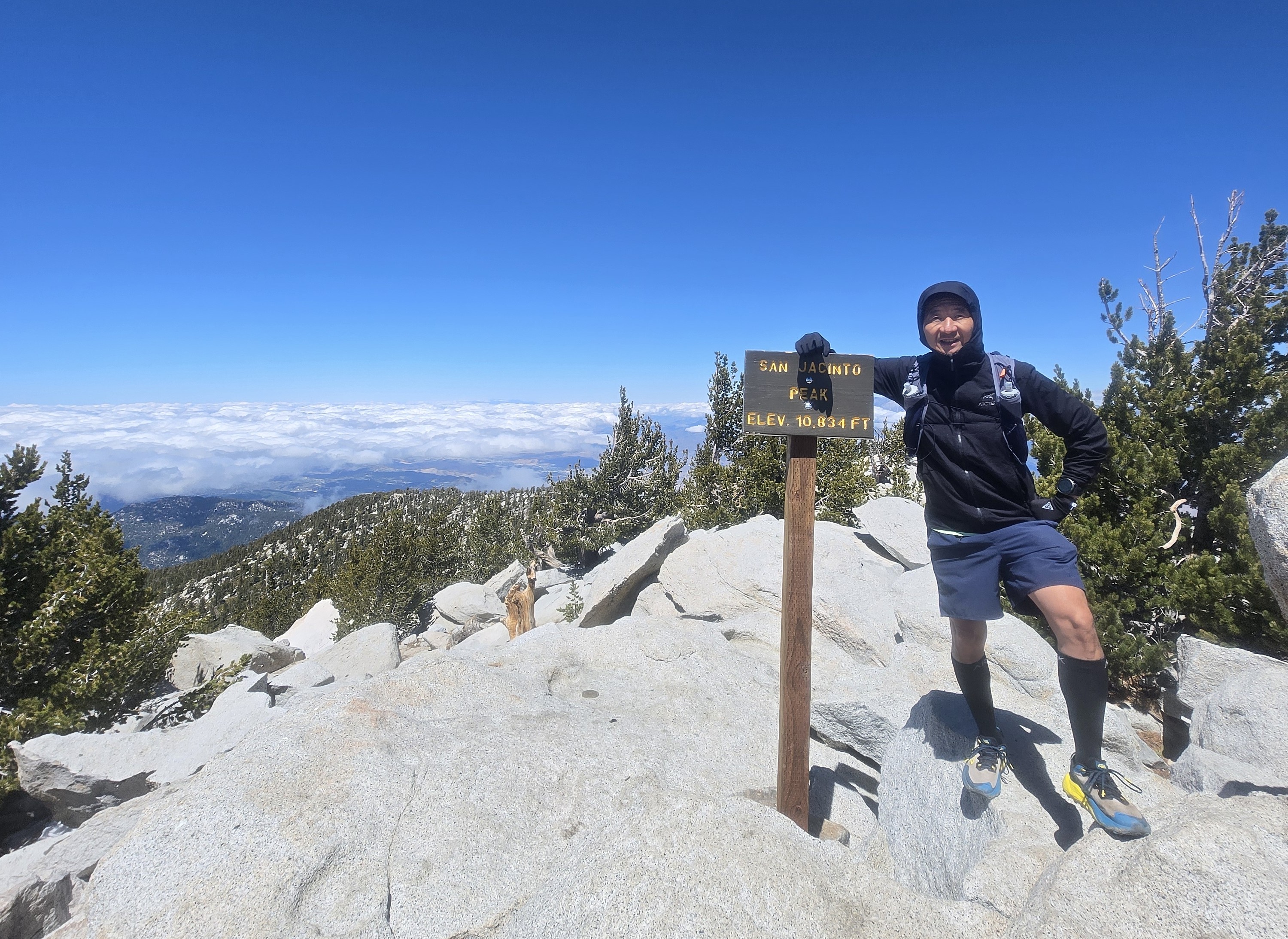 Lone hiker summits San Jacinto Peak, amongst a sea of clouds in the background.