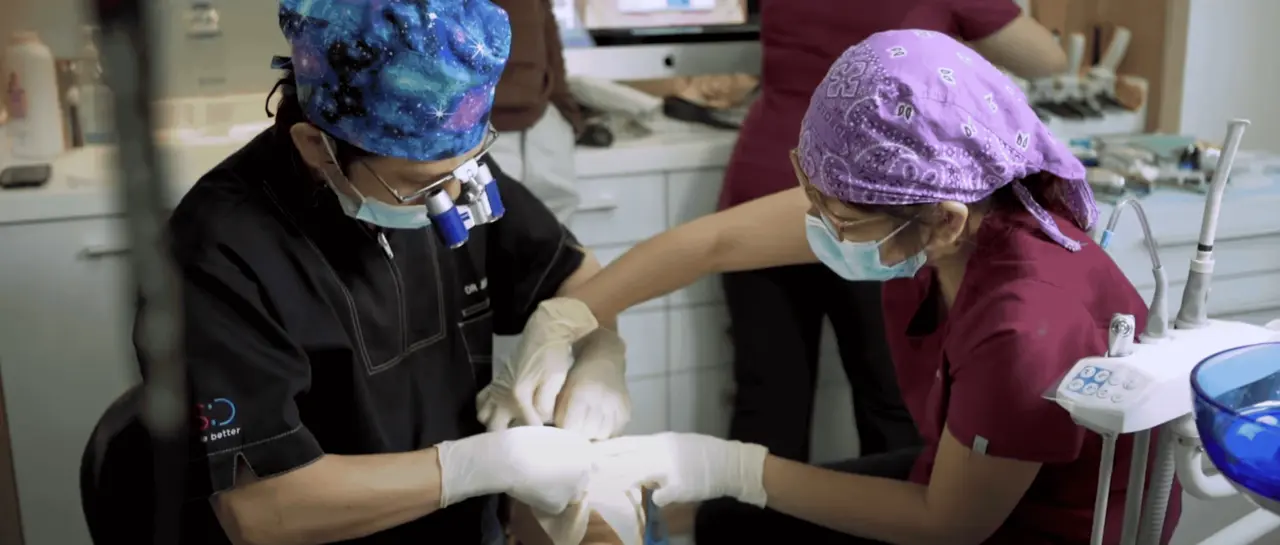 Two dental professionals wearing masks and gloves performing a dental procedure on a patient in a clinic.