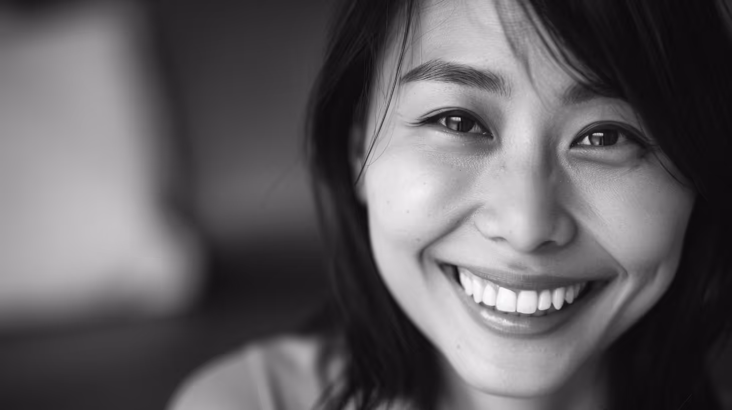 Young woman with long dark hair smiling brightly, resting her face on her hand.