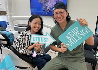 Smiling female patient sitting in a dental chair next to a male dentist holding signs that say 'DENTIST' and 'KEEP SMILING!'