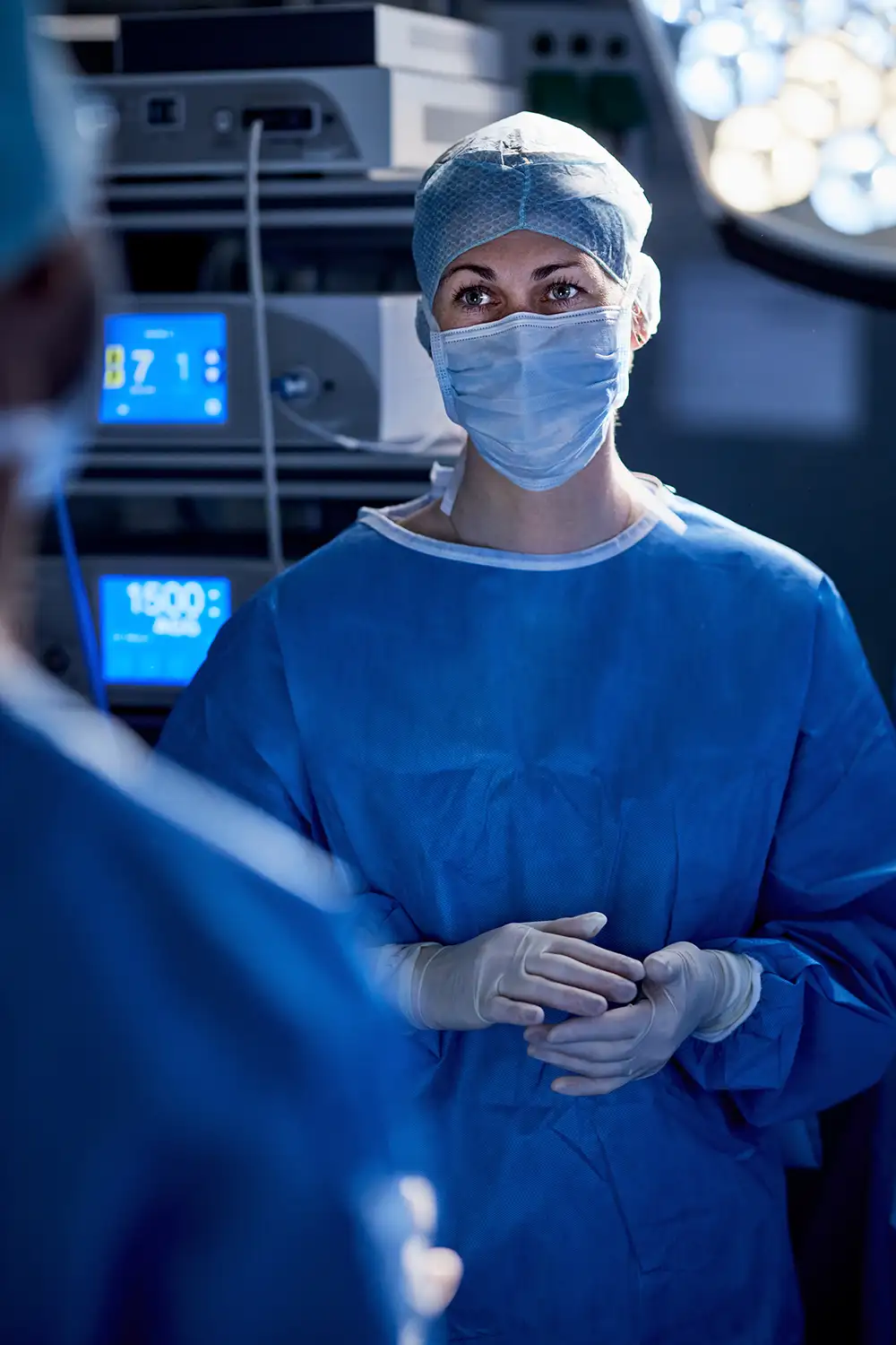 Surgeon looks at another surgeon in an operating room. Monitors showing vital signs are illuminated behind her.