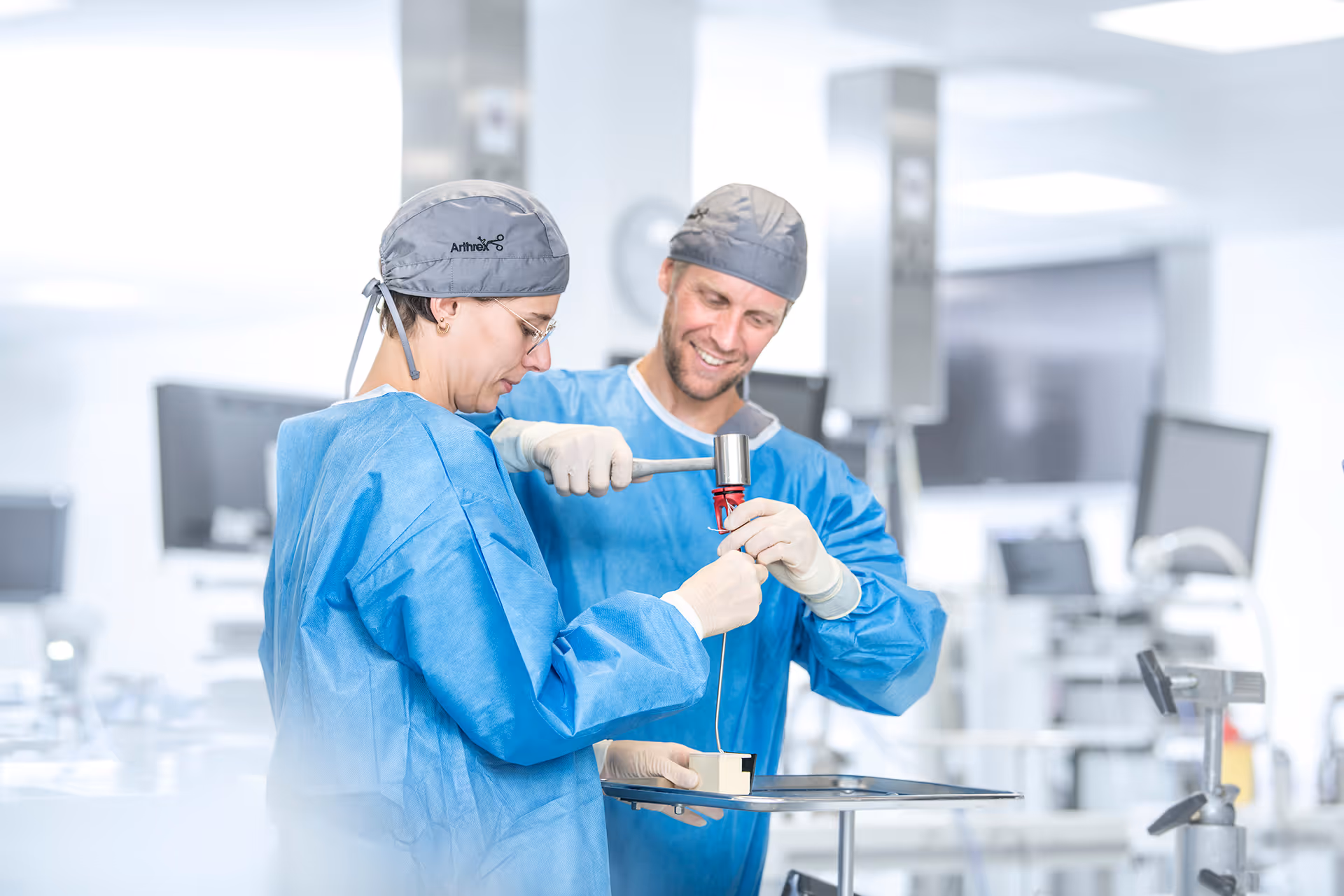 Two surgeons train in a lab. One holds a suture delivery device while the other impacts it with a mallet.