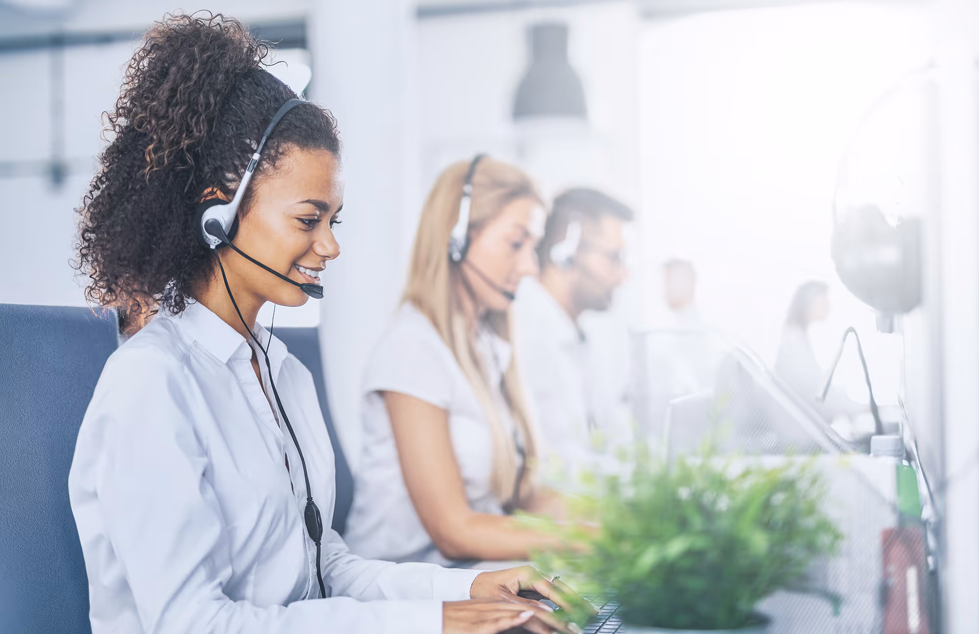 Two women and one man in a customer care call center, with the smiling woman in the foreground in focus and the man blurred in the background.