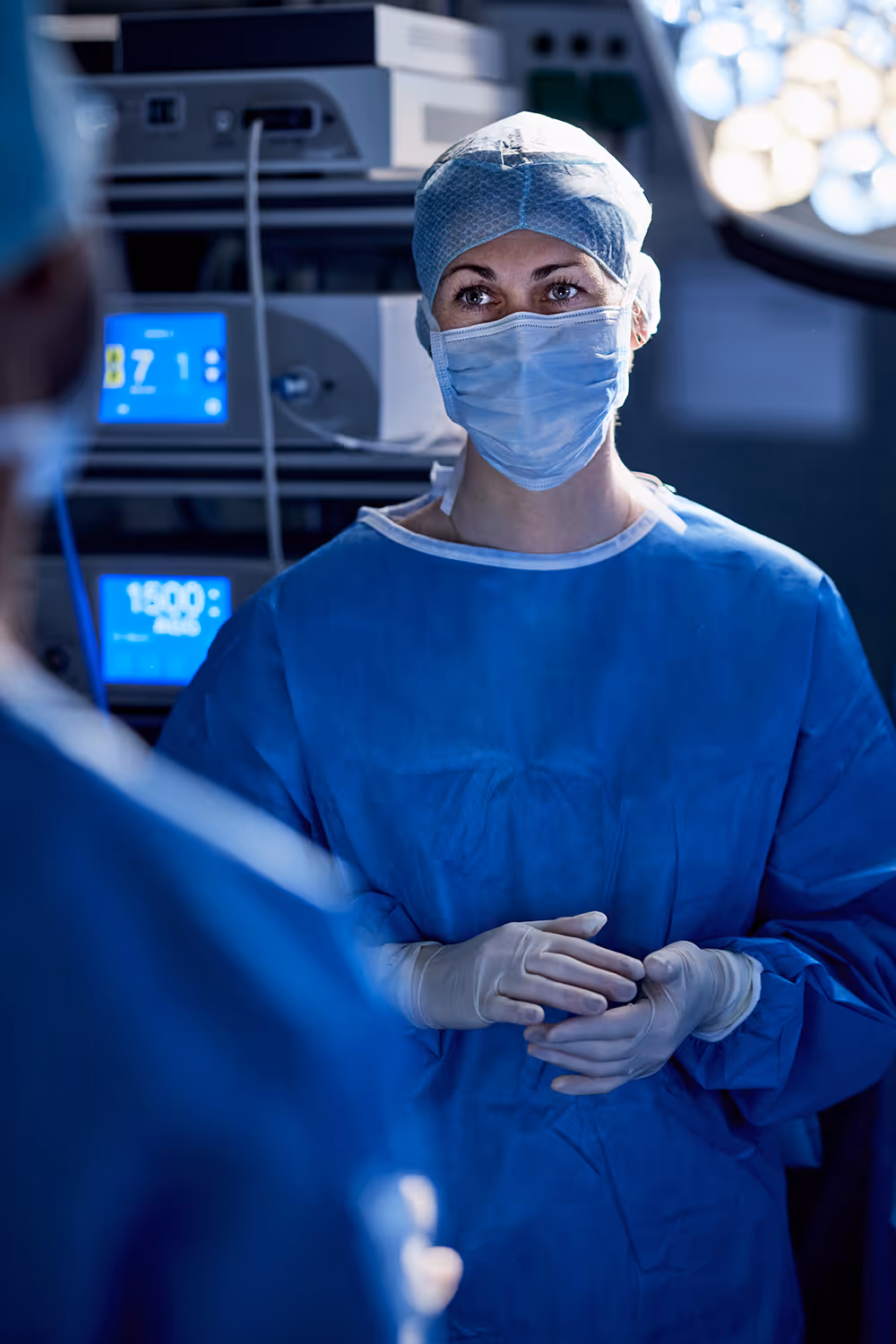 Surgeon looks at another surgeon in an operating room. Monitors showing vital signs are illuminated behind her.