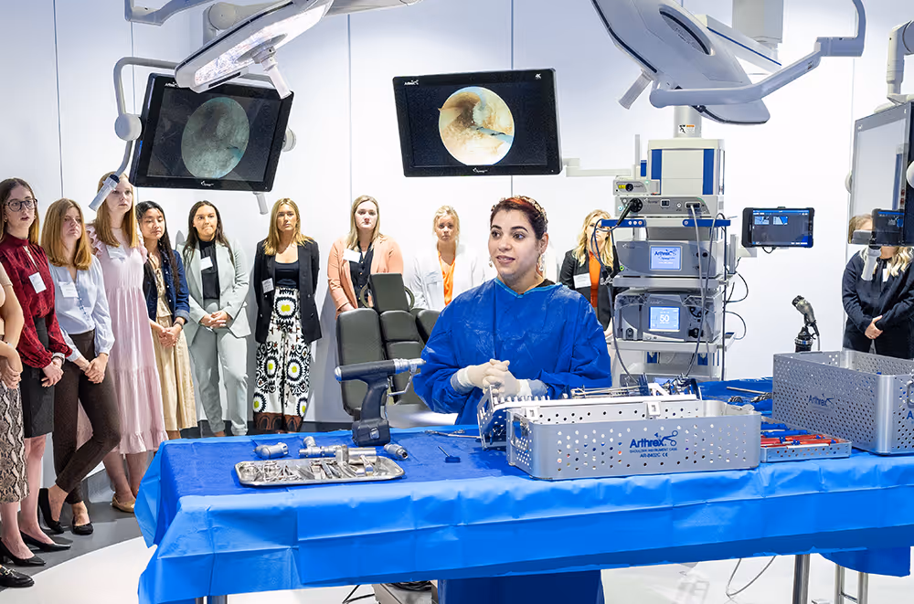 Medical education session where participants stand in a semicircle, observing an instructor as she explains the uses of various tools laid out before her.
