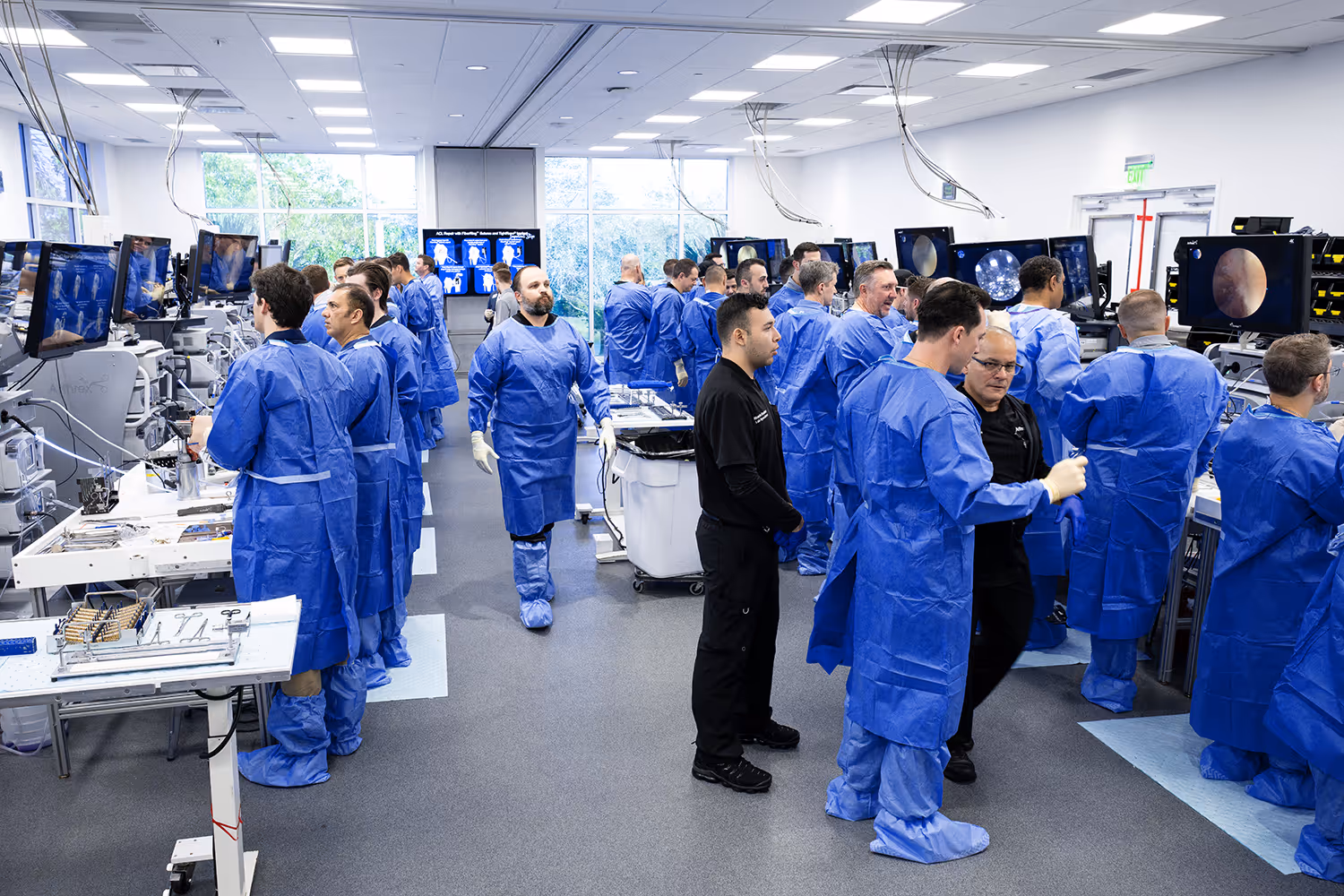 Surgeons wearing scrubs training in a brightly lit surgical lab. Instructor walks down the aisle reviewing the hands-on work of student surgeons.