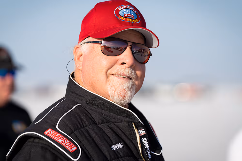 A man in sunglasses, a baseball cap, and a racing jacket stands outside.