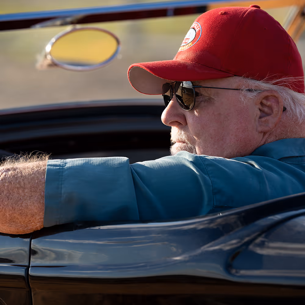 A man in sunglasses and a baseball cap drives a convertible car with the top down.