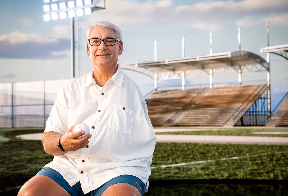 A one-armed man holding a baseball in his right hand sits near a baseball field.