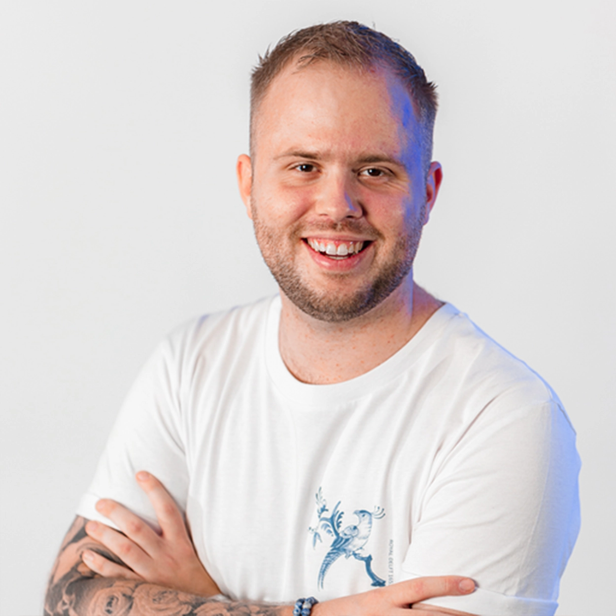 Lachende man met kort haar, baard en tatoeages op zijn onderarmen, gekleed in een wit t-shirt met blauwe vogelprint.