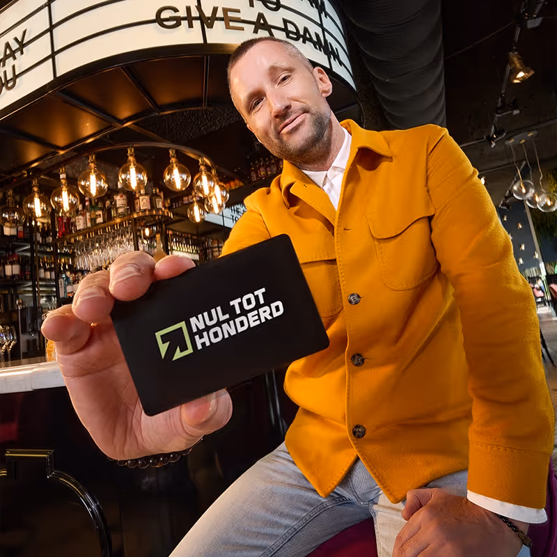 Man in een oranje jas toont een kaart met het logo en de tekst 'Nul tot honderd' in een bar.