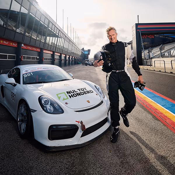 Man in racepakker poseert naast een witte raceauto met tekst 'Nul tot Honderd' op het circuit.