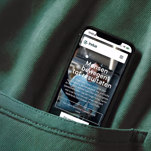 Smartphone met een website van Indus met Nederlandse tekst over impact en samenwerking, in een groene broekzak.