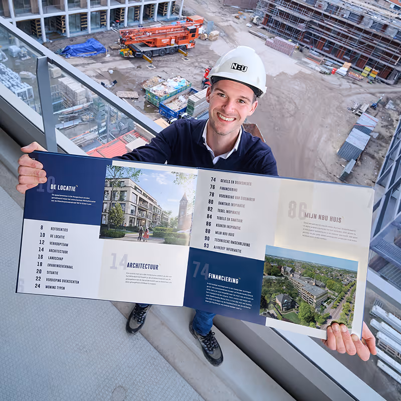 Vrolijke man met een witte bouwhelm die een open brochure over architectuur en financiering toont op een bouwplaats.