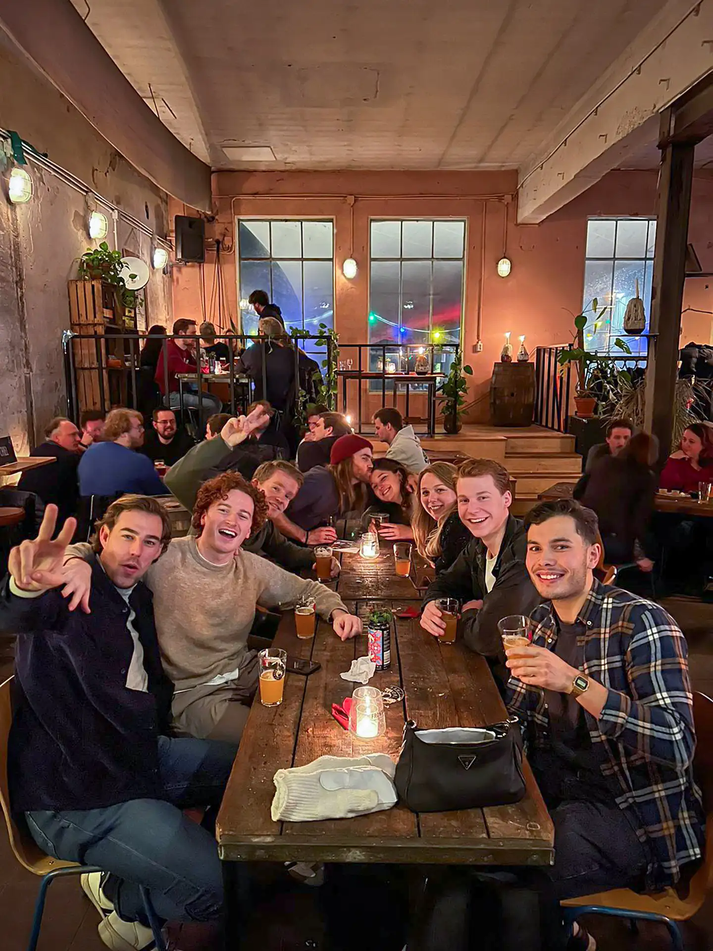 Een groep jonge mensen zit samen aan een houten tafel in een gezellige, sfeervol verlichte bar, drinkt bier en lacht naar de camera.