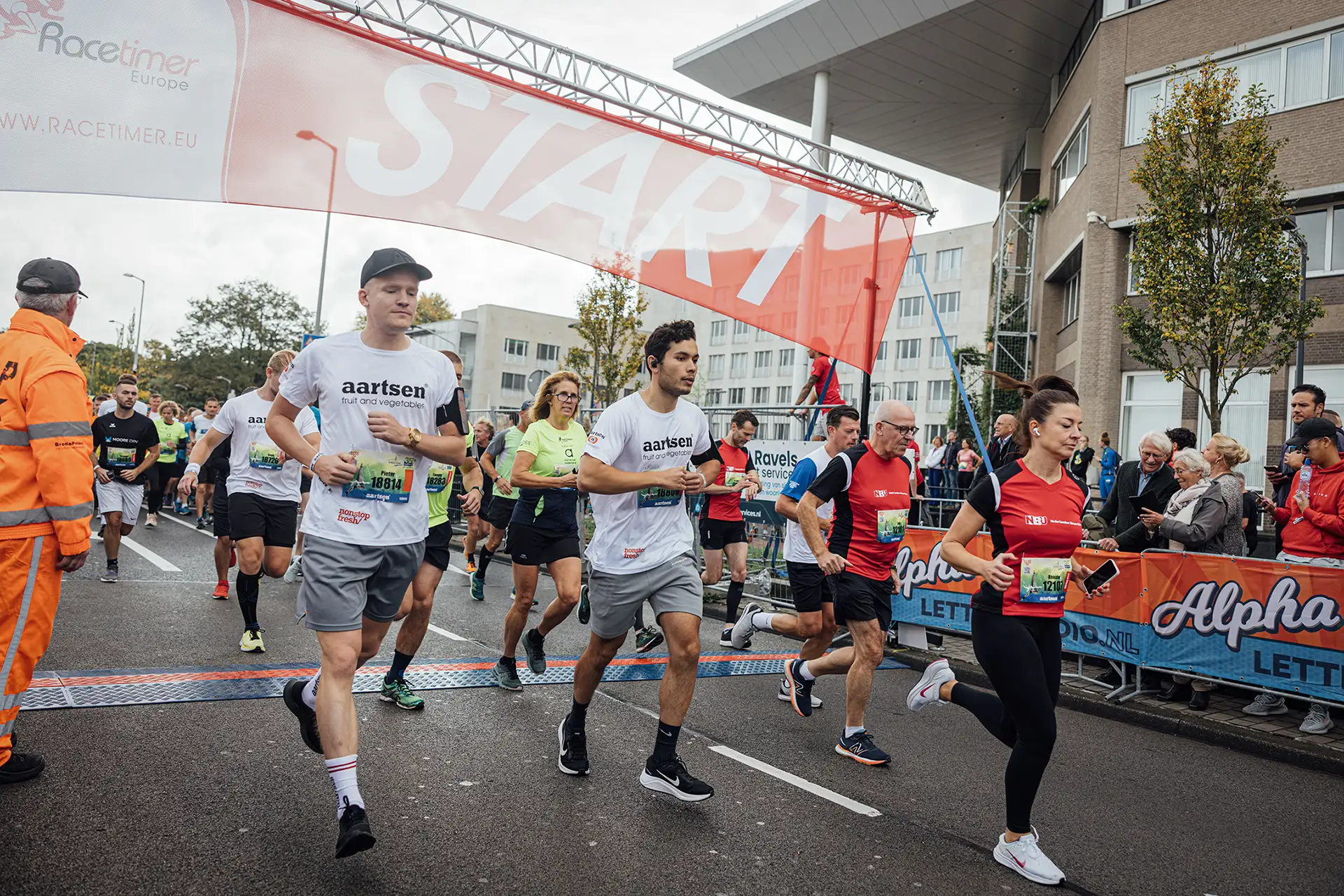 Groep hardlopers die een straatrace starten onder een grote START-boog met toeschouwers langs de route.
