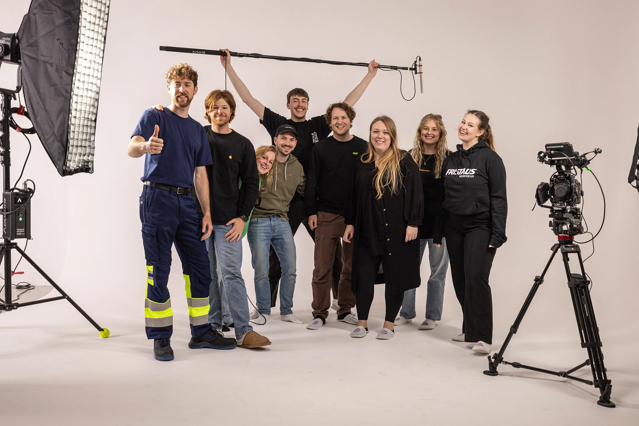 Groep van negen mensen in een fotostudio met professionele uitrusting, lachend in casual kleding en één persoon geeft een duim omhoog.