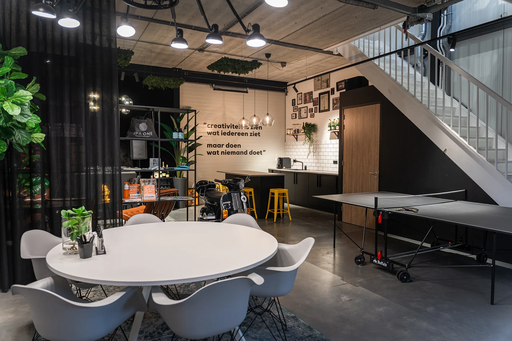 Moderne kantoorruimte met witte ronde tafel en stoelen, pingpongtafel, scooter, en keukenhoek met gele krukken onder een trap.