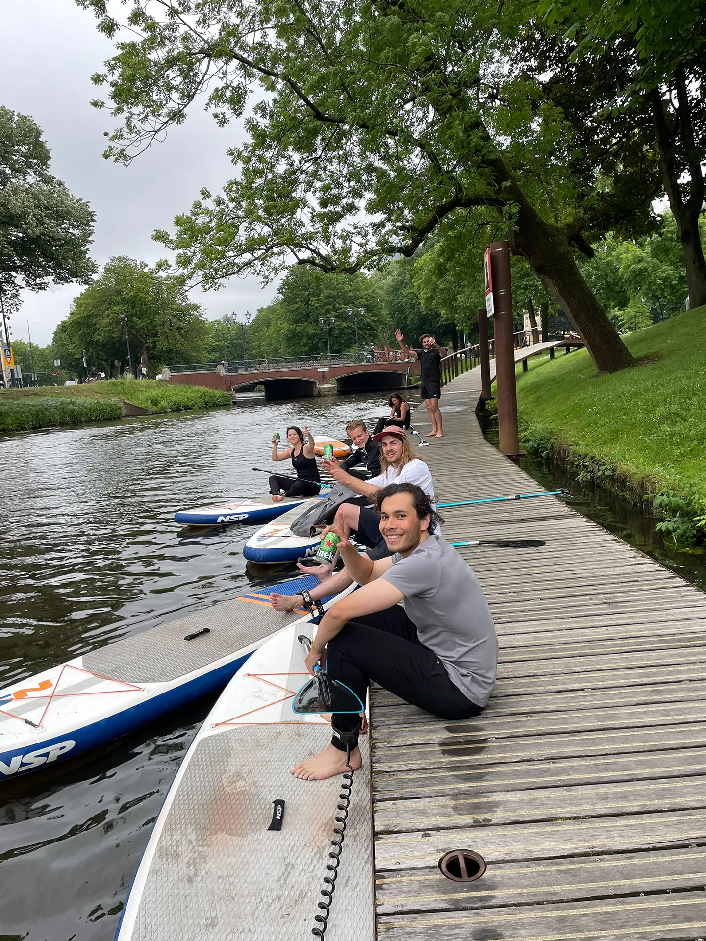 Vijf mensen zitten op paddleboards langs een houten steiger langs een kanaal, genietend en zwaaiend met blikjes in hun handen.