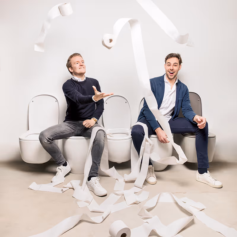 Twee mannen zitten op naast elkaar geplaatste toiletten met rollen wc-papier die door de lucht vliegen.