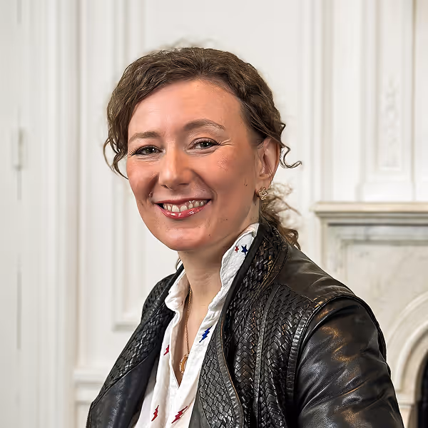 Femme souriante aux cheveux bouclés portant une chemise blanche et une veste en cuir noir dans un intérieur clair.