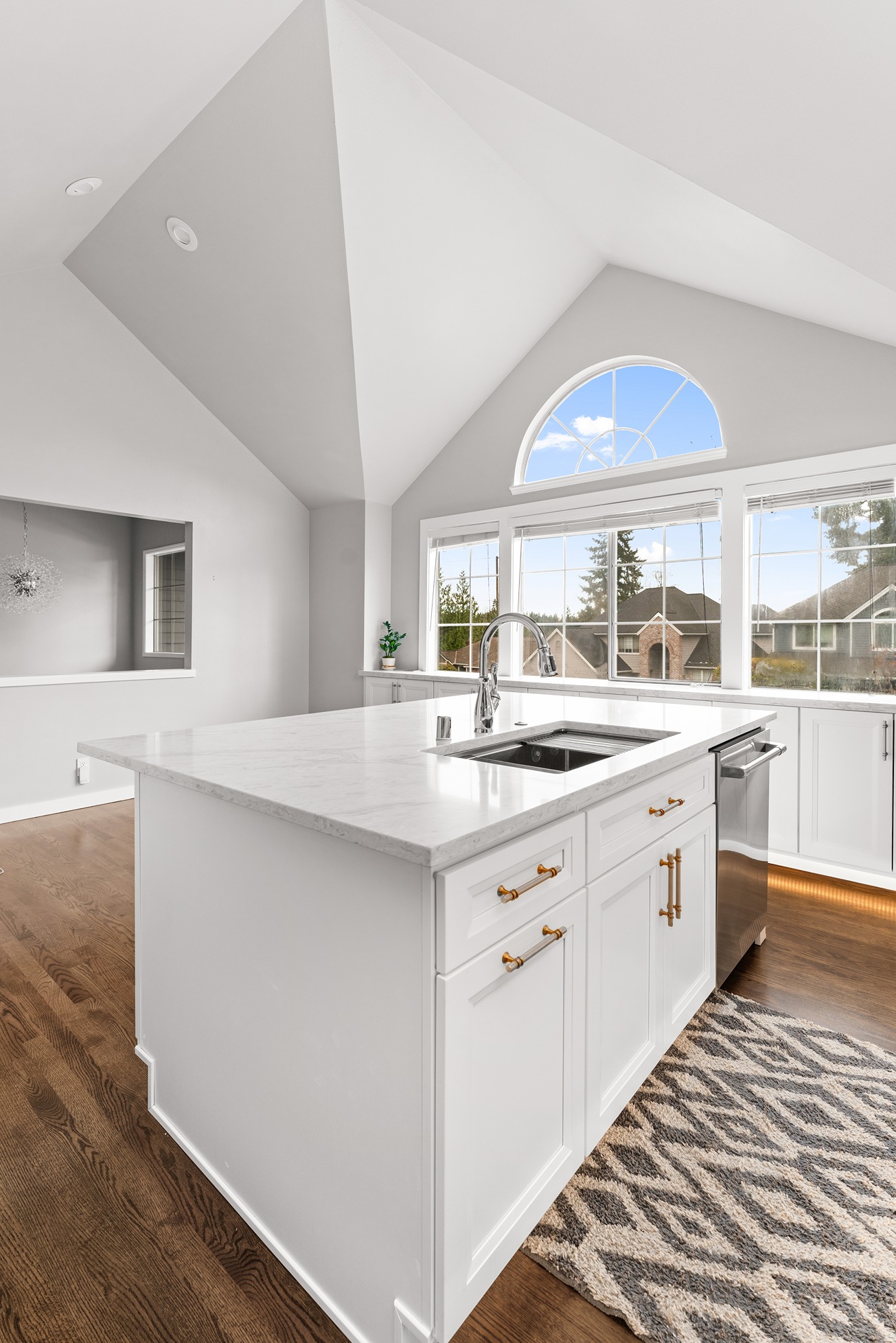 Modern kitchen with a vaulted ceiling and large arched window, featuring a central island with white cabinetry, marble countertops, brass hardware