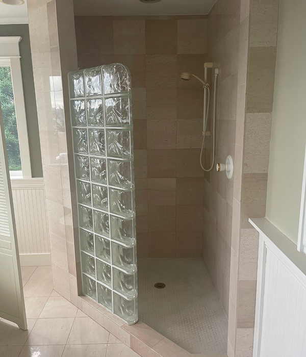 Outdated walk-in shower with beige tile walls and floor, featuring a partial glass block wall, a basic handheld showerhead, and minimal lighting