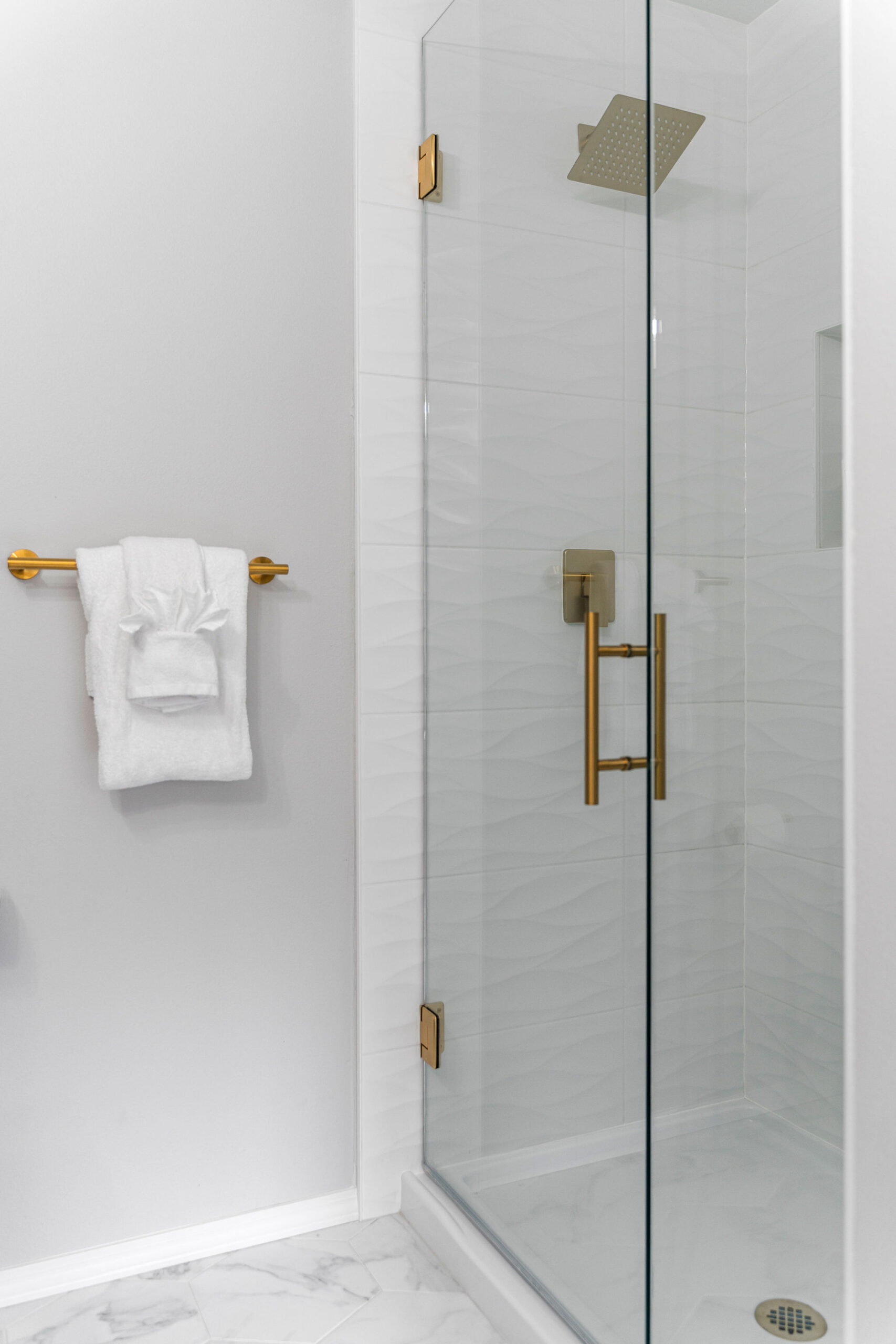 Modern glass shower with gold hardware, featuring a rain showerhead, minimalist wave-textured white tile walls