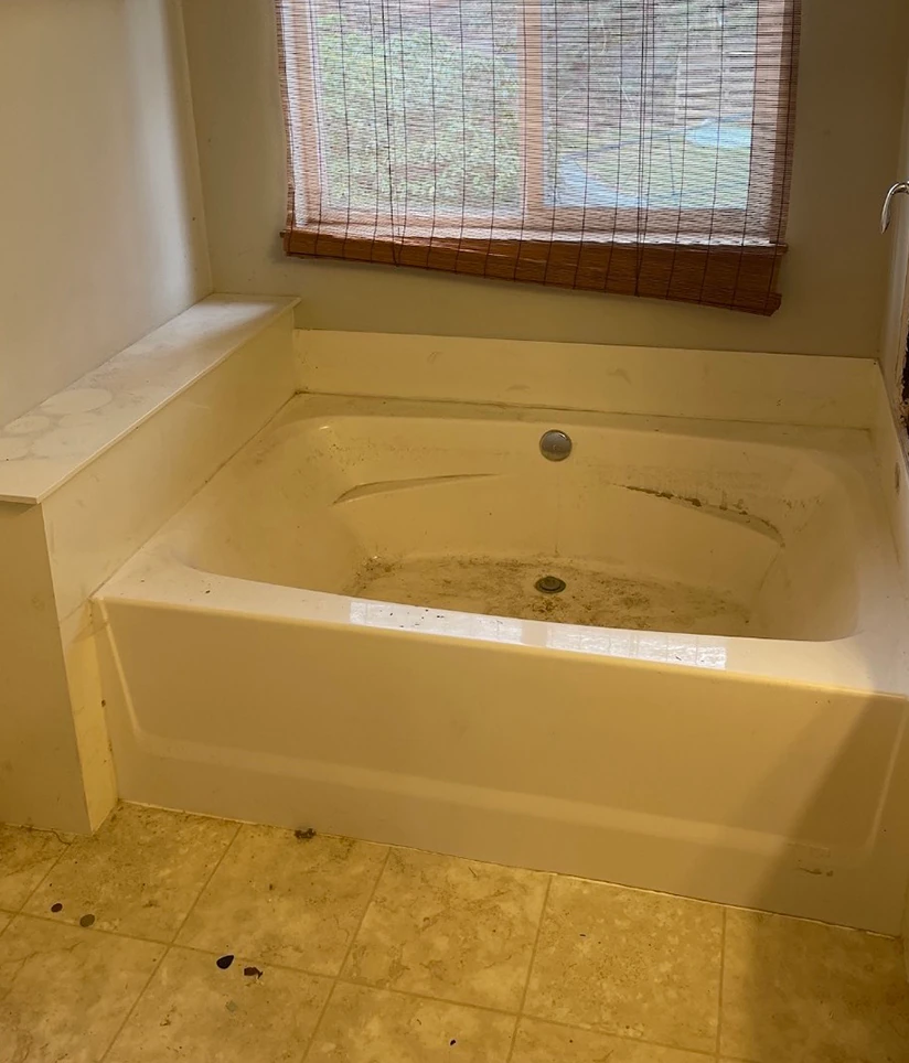 an old, built-in, stained bathtub with beige tile flooring, bamboo window shade, and dated finishes