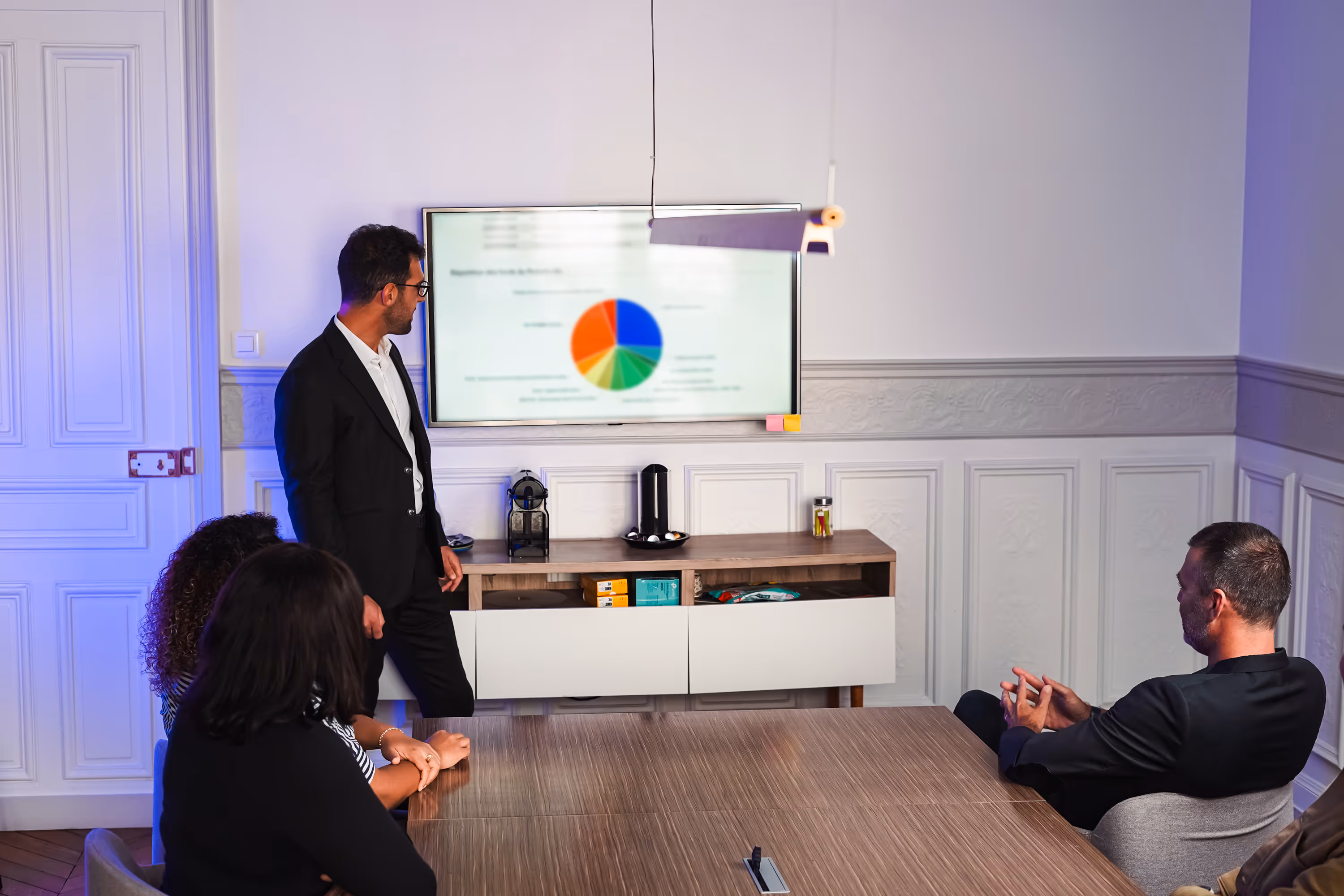 Man in a suit giving a presentation to three seated colleagues in a meeting room with a pie chart displayed on a wall-mounted screen.