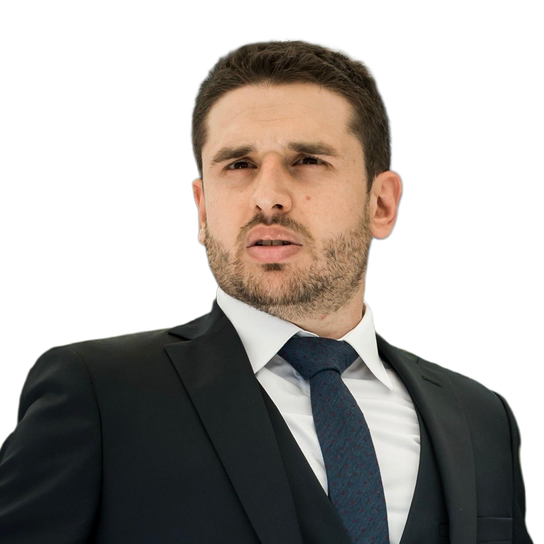 Serious man with short dark hair and beard wearing a black suit and blue patterned tie outdoors.