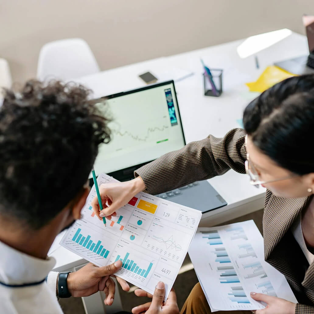Two people in an office reviewing financial documents with charts and graphs. One is pointing at a paper with a pen. A laptop displaying a line graph is open on the table.