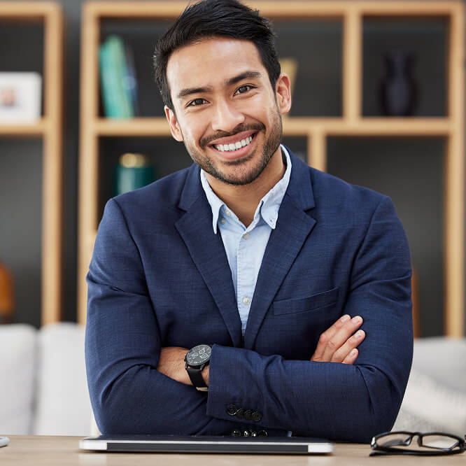 "A man in a blue blazer smiles with arms crossed, seated at a desk with a closed laptop and glasses. Shelves with books and decor are in the blurred background."
