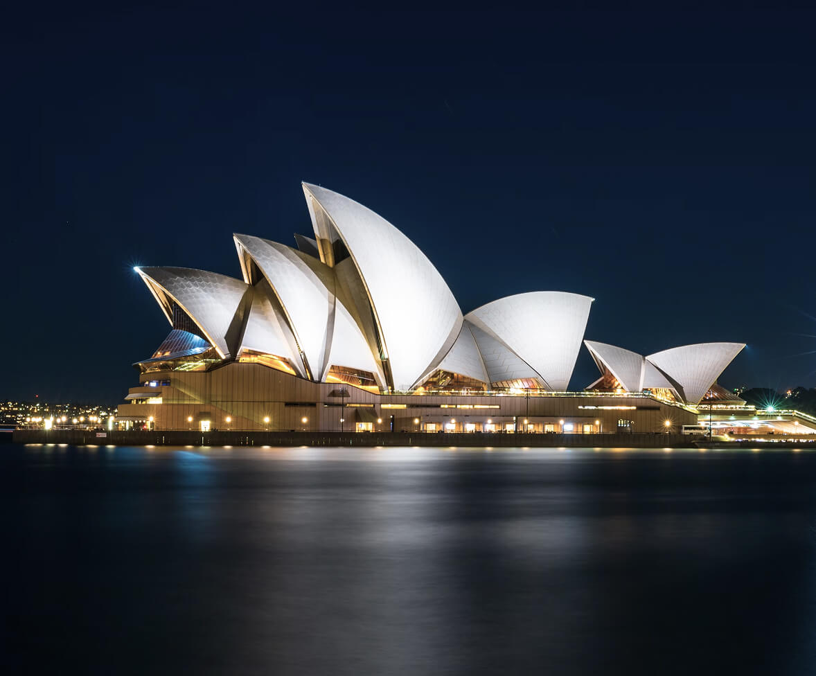 "Sydney Opera House illuminated at night, with white sail-like roofs reflecting on the calm harbor water under a dark sky."